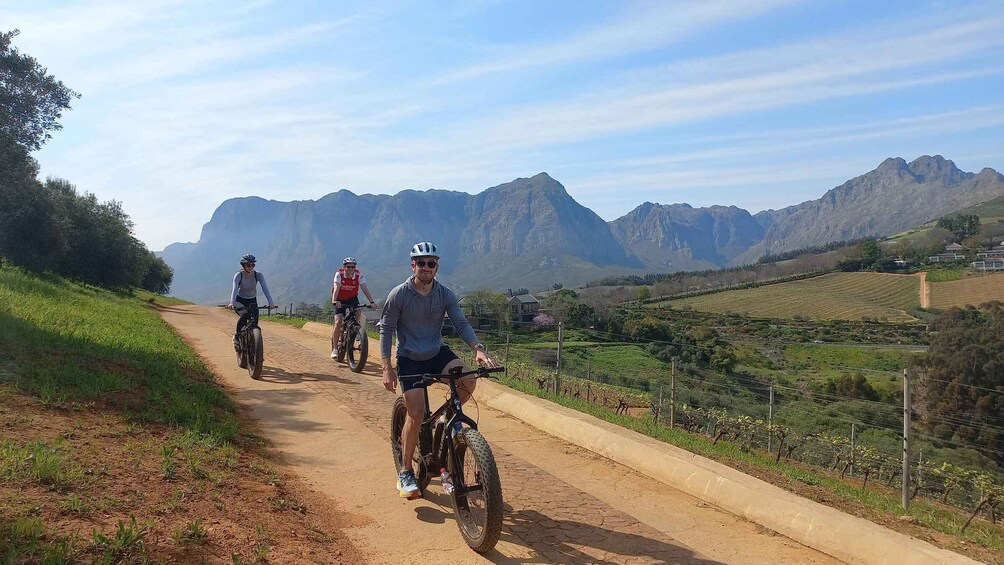 Three cyclists on a dirt path with mountains in the background. 1