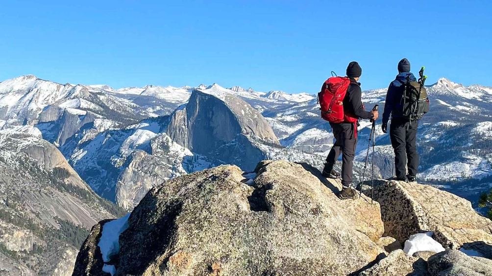 Two hikers on a rocky mountain peak with a snowy mountain range in the background. 1