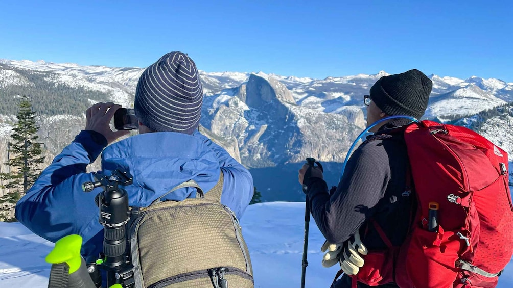 Two people with backpacks and cameras, overlooking a snow-covered mountain range. 3