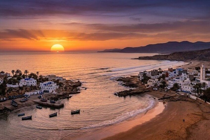 A coastal village at sunset with boats, buildings, and a beach. 1