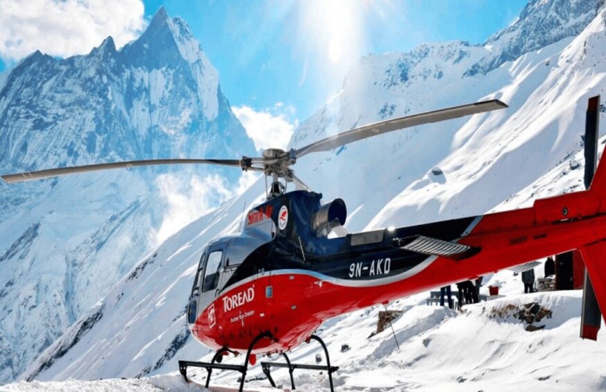 A red and black helicopter with 'Toread' and '9N-AKO' on its side, hovering over a snowy mountain landscape with a clear sky. 2