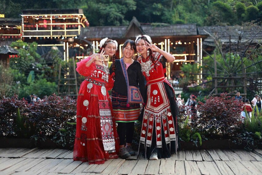 Three people in traditional red attire posing in front of a wooden structure with illuminated decorations. 1