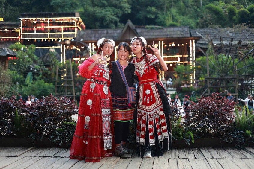 Three people in traditional attire standing on a wooden deck with a backdrop of illuminated structures and greenery. 3