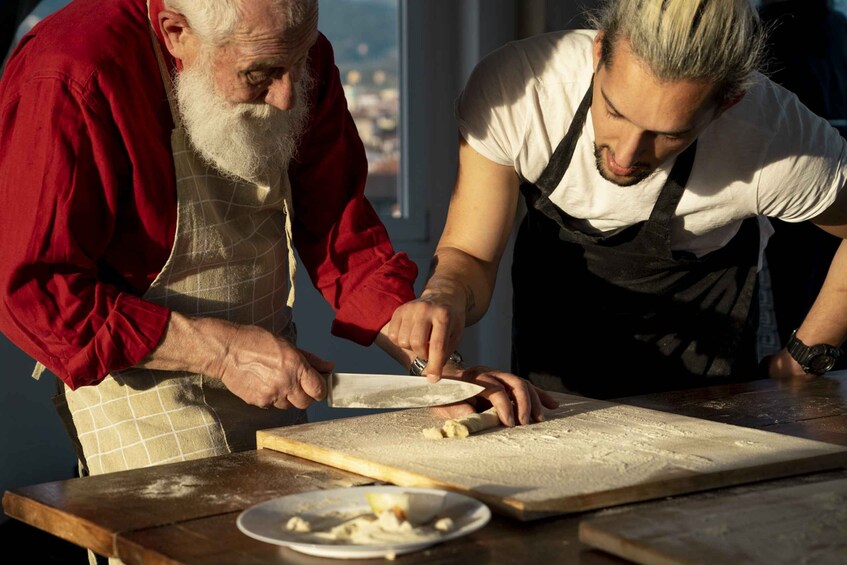 Twee personen bereiden deeg op een houten tafel, met een deegroller en een kom vol ingrediënten. 3