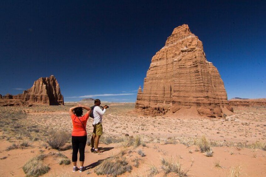 A desert landscape with a prominent rock formation and two people taking photographs. 2