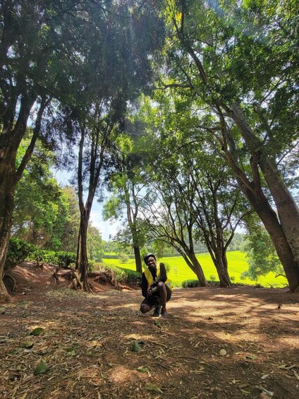 A person squatting in a forested area with trees and a green field in the background. 4