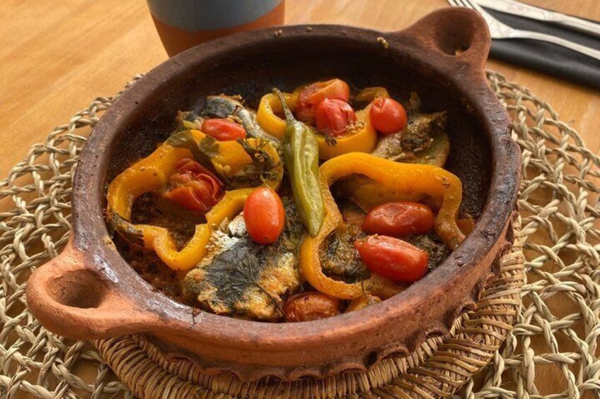 A clay pot with a stew containing fish, bell peppers, and tomatoes. 3