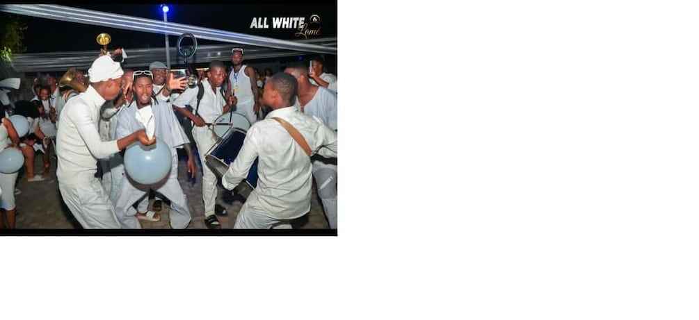 A group of people in white attire, some holding balloons, gathered in a semi-outdoor setting with a tent in the background. 5