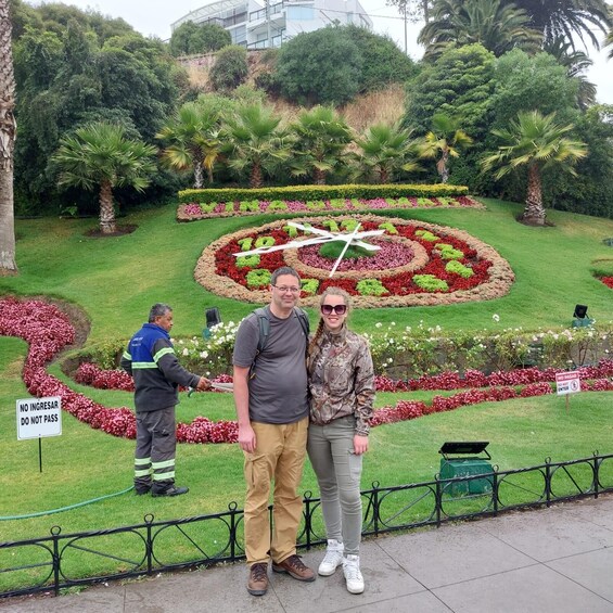 A man and a woman standing in a garden with a circular flower bed and a white cross in the center. 2