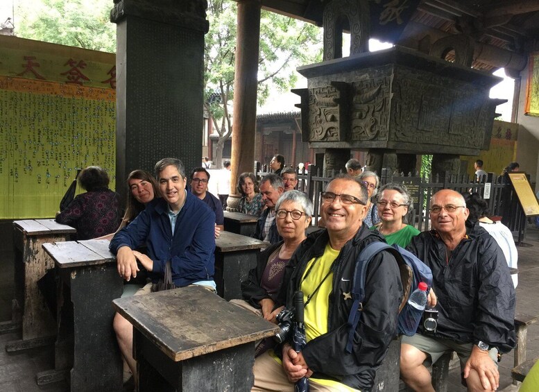 A group of people seated at wooden tables under a covered structure with stone carvings, surrounded by trees. 5