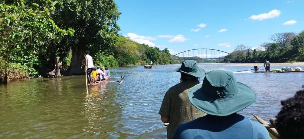 A river scene with people on boats, a bridge in the background, and lush greenery along the banks. 3