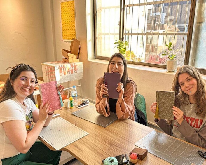 Three people holding notebooks at a wooden table with crafting materials. 3