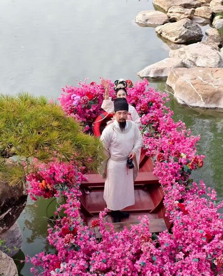 A person in traditional attire standing on a boat surrounded by pink flowers. 3