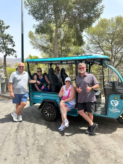 A group of people in a turquoise golf cart with 'SVIWO' branding, parked outdoors with trees and a clear sky in the background. 3