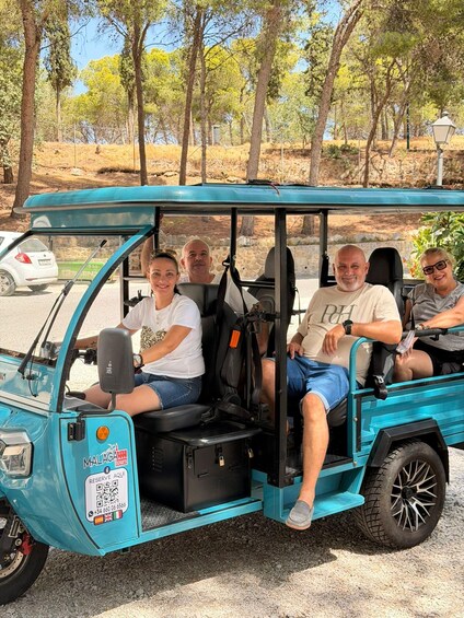A turquoise golf cart with four passengers, parked on a gravel path with trees in the background. 2