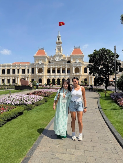 A historic building with a flag, two people standing in front, and a flower garden. 1