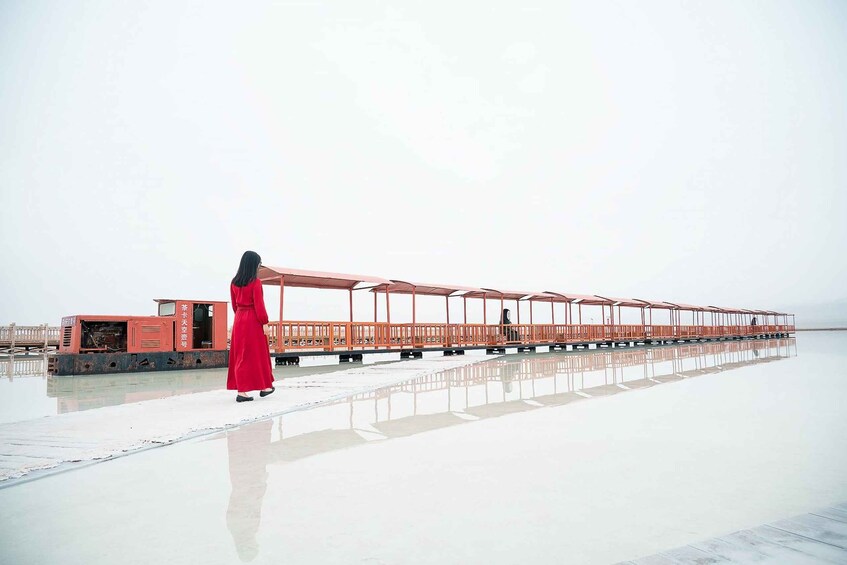 A person in a red dress walking on a reflective water surface with a row of red canopies and a stationary train car in the background. 2