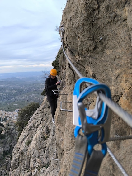 A person rappelling down a rocky cliff with a harness and helmet, overlooking a valley with buildings and a winding road. 5