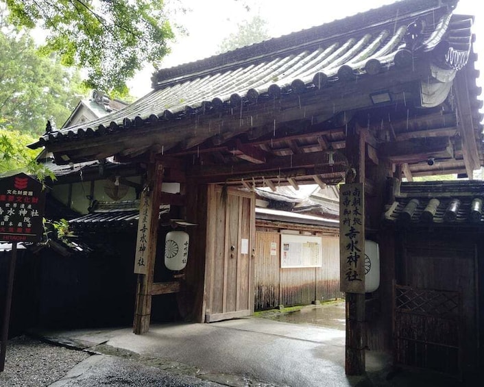 A traditional Japanese gate with wooden pillars and a tiled roof, leading to a building with a signboard. 3
