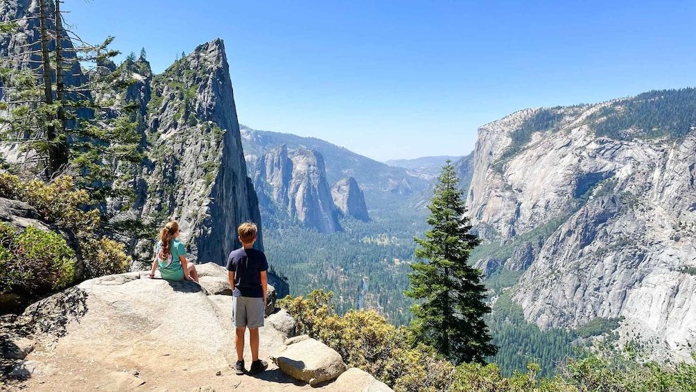 Two people standing on a rocky trail overlooking a canyon with steep cliffs and dense forest. 4