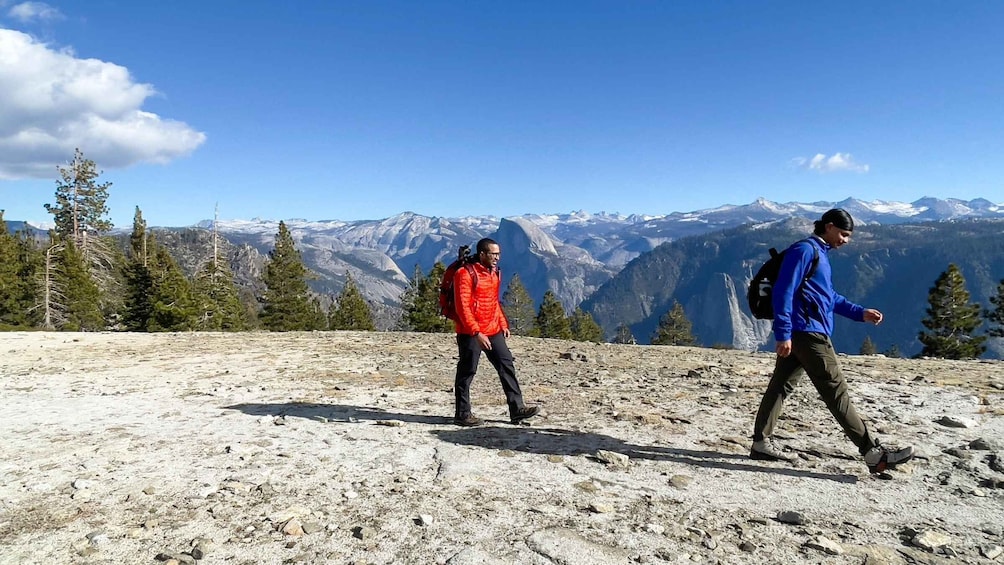 Two hikers on a rocky mountain trail with snow-capped mountains in the background. 3