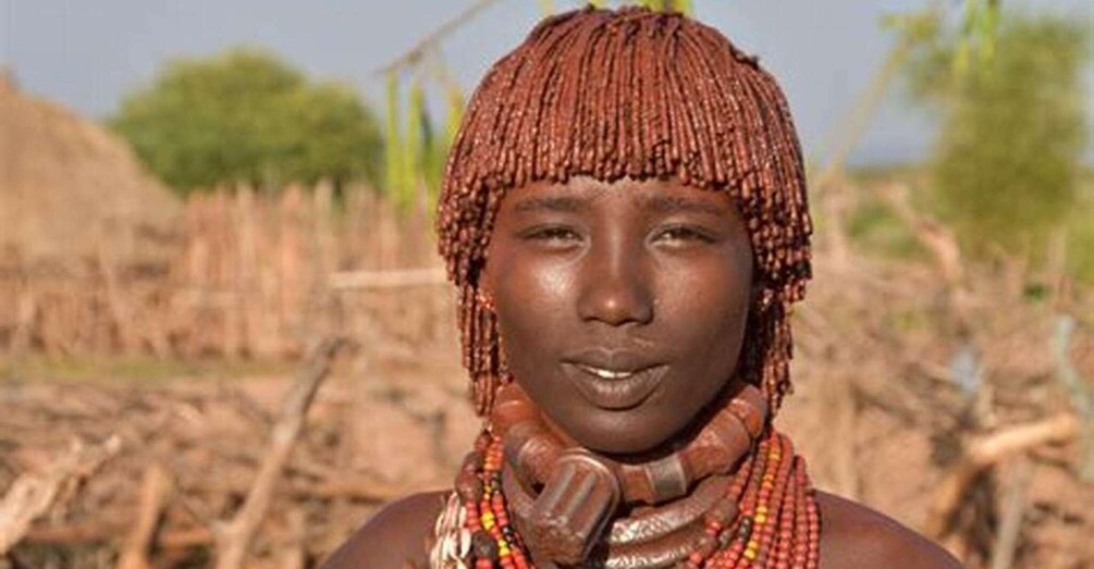 A person with braided hair and beaded jewelry in a rural setting with trees and wooden structures. 2