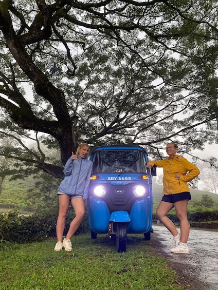 Two people standing beside a blue vehicle with headlights on, surrounded by trees and greenery. 5