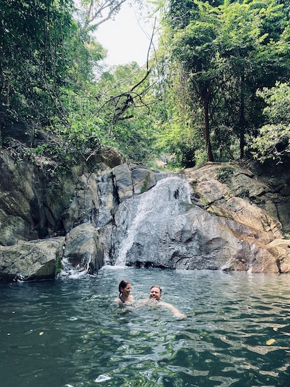 A waterfall in a forested area with two people swimming in the water. 4