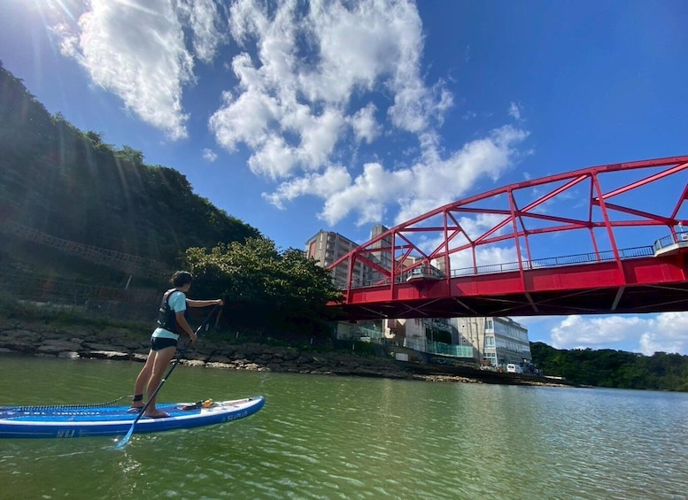 A person paddleboarding on a river with a red bridge in the background. 1