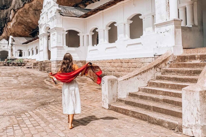 A person in a white dress with a red scarf walking up stone steps towards a white building with arches. 5