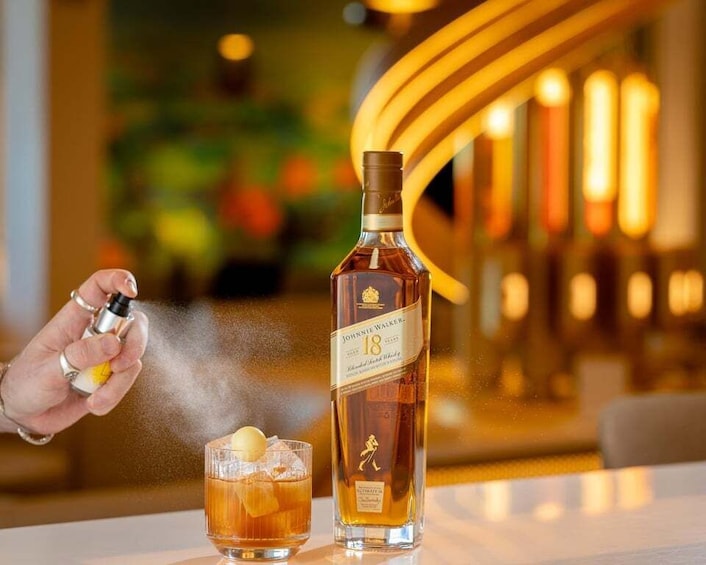 A hand spraying a mist over a glass of whiskey and a bottle labeled 'Johnnie Walker 18' on a bar counter. 2