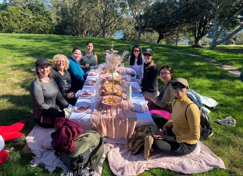 A group of people sitting around a picnic table with food and drinks in a grassy park. 2