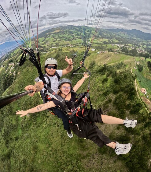 Two people paragliding over a lush green landscape with rolling hills and a cloudy sky. 1