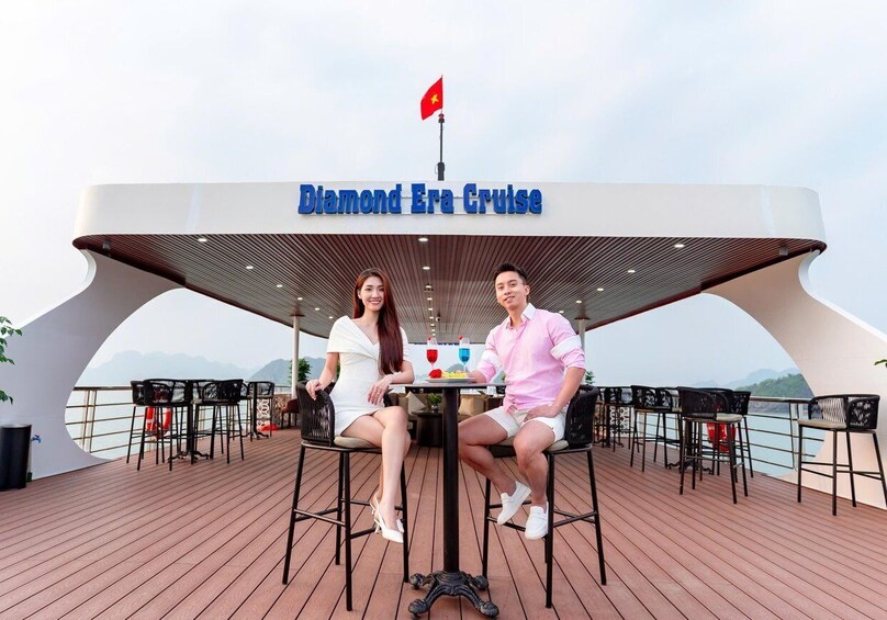 A couple seated at a table on a cruise deck with a sign reading 'Diamond Era Cruise' and a flag in the background. 3
