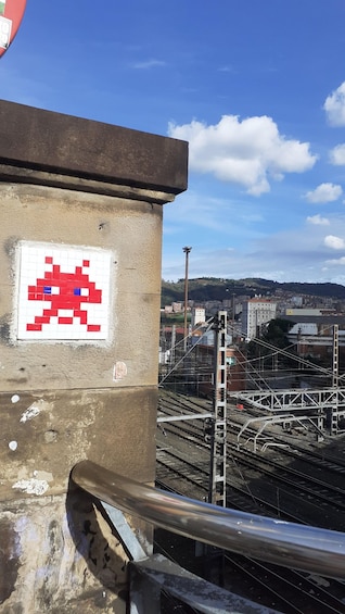 A pixelated alien on a wall, a metal railing, and a cityscape with trains in the background. 1