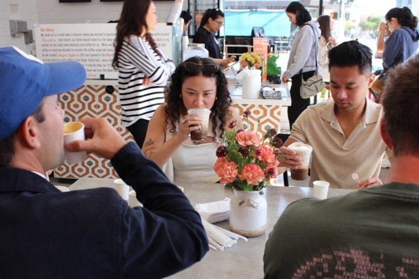 Des personnes sont assises à une table dans un café, devant des boissons, un vase de fleurs et un mur décoré de motifs. 1