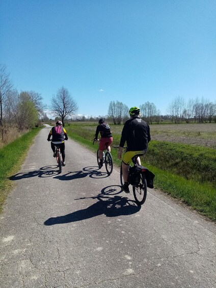 Three cyclists on a paved path with a rural landscape, including trees and open fields. 3