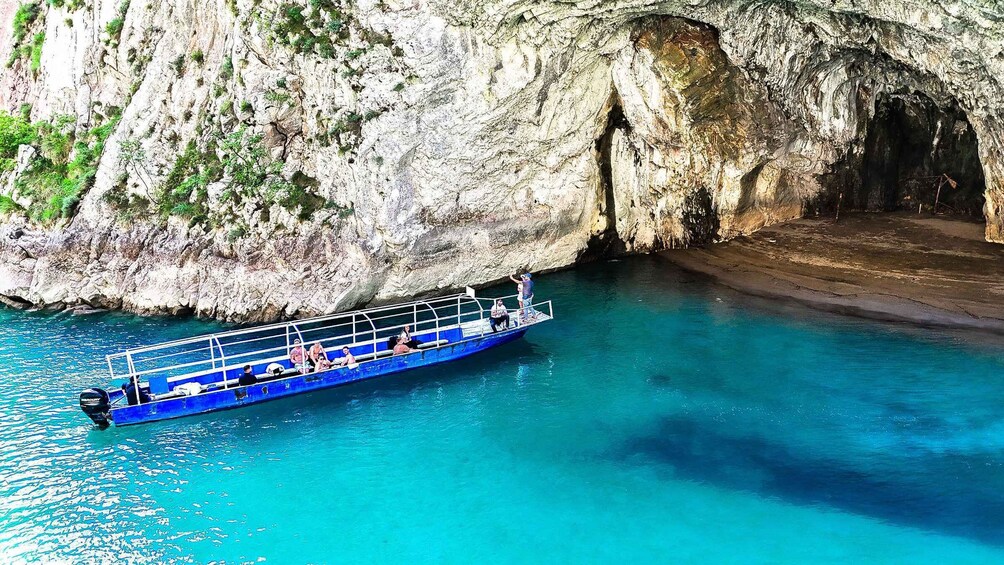 A blue boat with passengers on a clear turquoise sea, near a rocky cliff with a cave entrance. 3