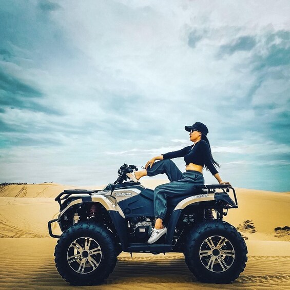 A person sitting on a quad bike in a desert with sand dunes in the background. 1