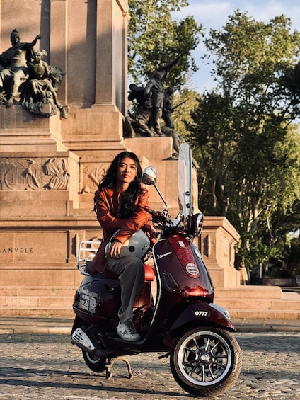 A person sitting on a red scooter with a helmet, in front of a stone monument with sculptures. 5