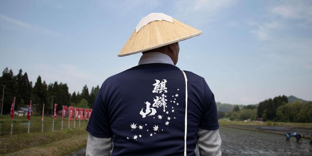 A person wearing a conical hat and a dark blue shirt with white text and symbols, standing by a river with trees and red flags in the background. 4