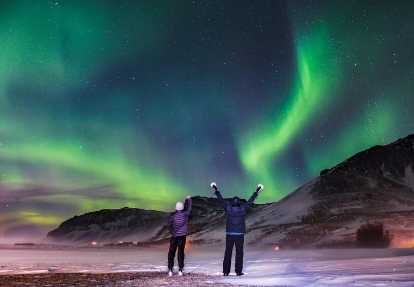 Två personer står på ett snötäckt landskap och blickar mot ett berg under en stjärnklar natthimmel, där norrskenet dansar. 1