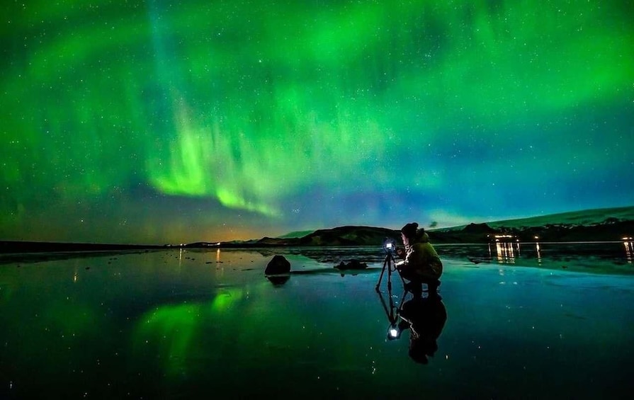 En person fotograferar norrskenet över en vattenyta som speglar ljuset, med berg i bakgrunden. 2