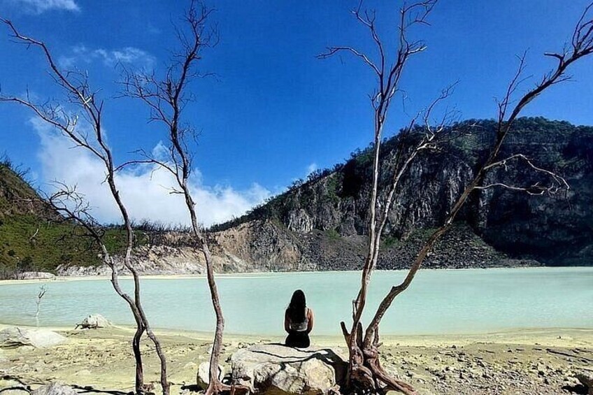 A person sitting on a rock by a calm lake with bare trees and a mountain in the background. 2