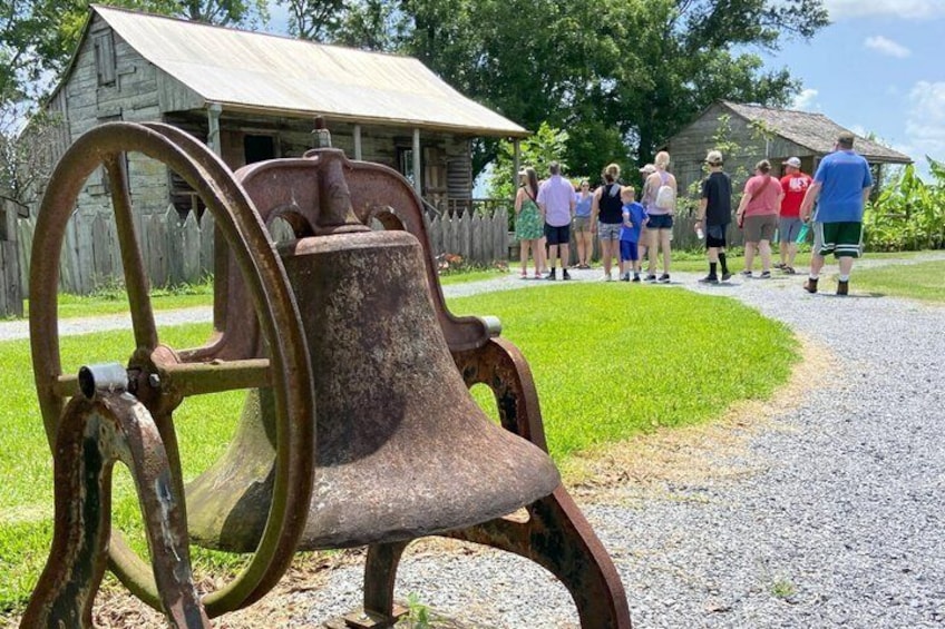 A rusty bell with a wheel, a gravel path, and a group of people standing in a grassy area with wooden buildings in the background. 2
