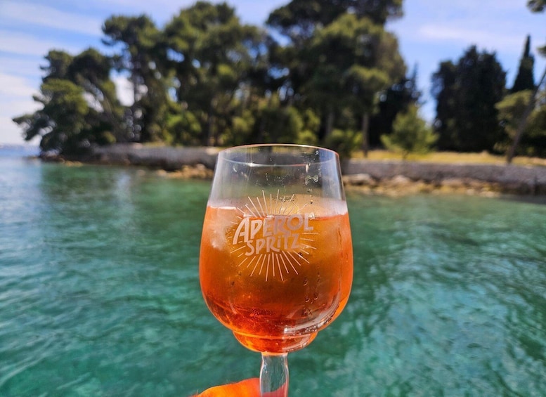 A glass of amber liquid with 'APEROL SPRITZ' on it, held against a backdrop of clear turquoise water and a tree-lined shore. 1