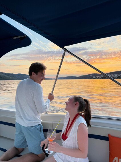 Two people on a boat, one holding a drink, with a sunset over water and hills in the background. 4