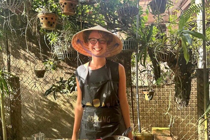Une personne portant un chapeau et un tablier, debout devant un jardin avec des plantes et des pots suspendus. 3