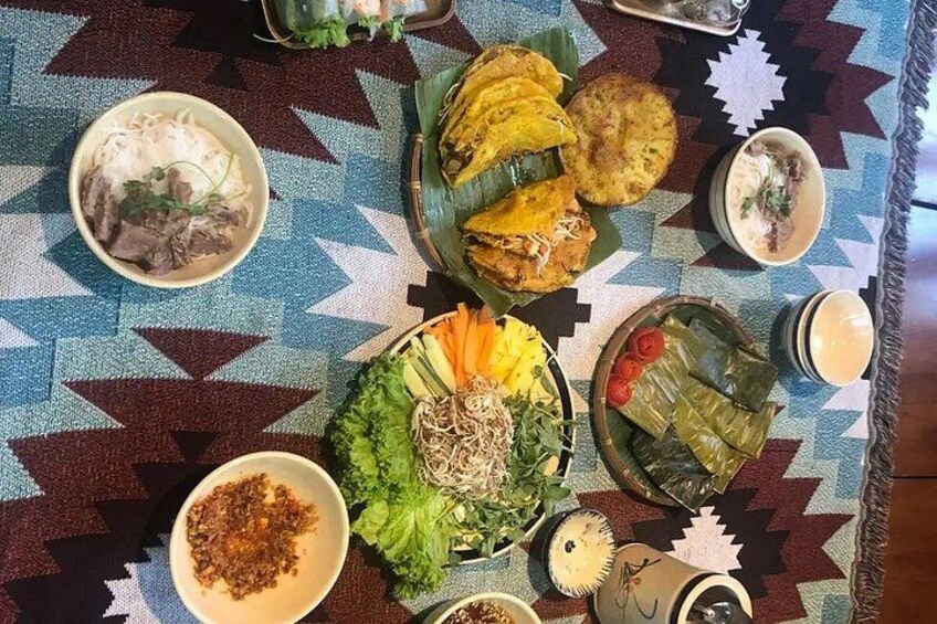 Une table dressée avec divers plats, notamment un bol de nouilles, une assiette de fritures et un plat de légumes enveloppés dans des feuilles. 2