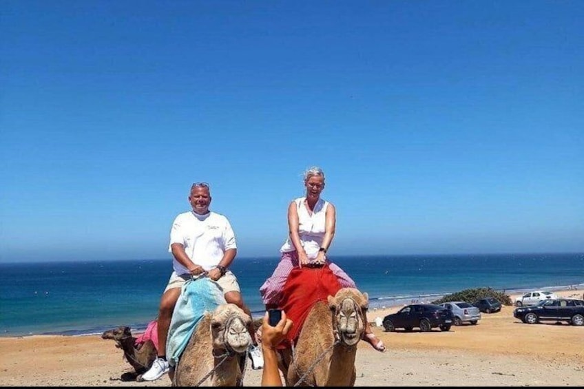 Two people riding camels on a beach with the ocean in the background. 2